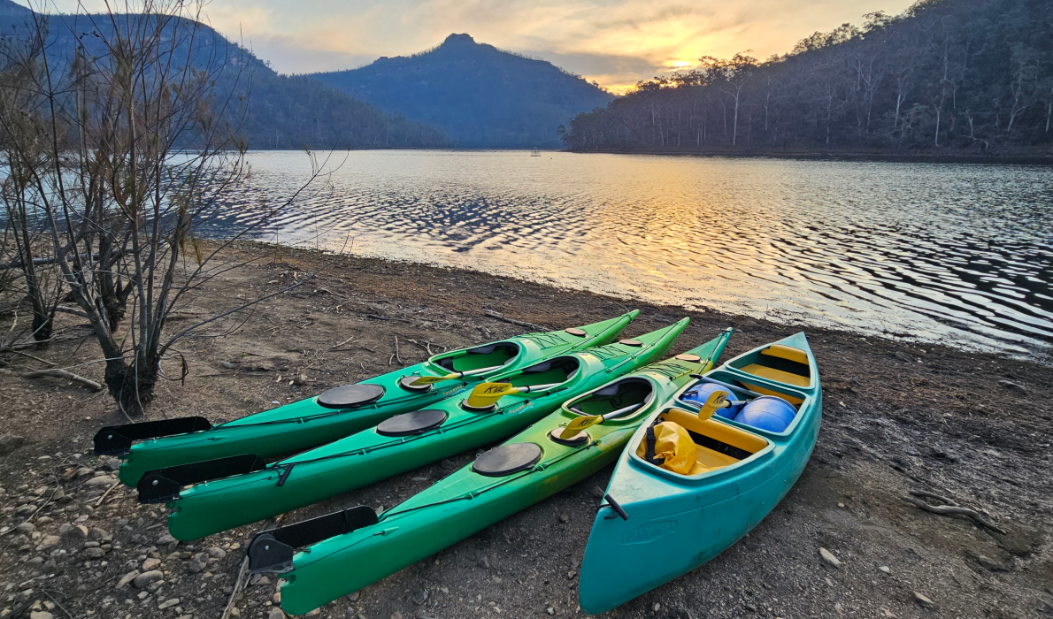 A peaceful image of canoes and kayaks lined up on the rivers edge with mountains and a sunset in the background