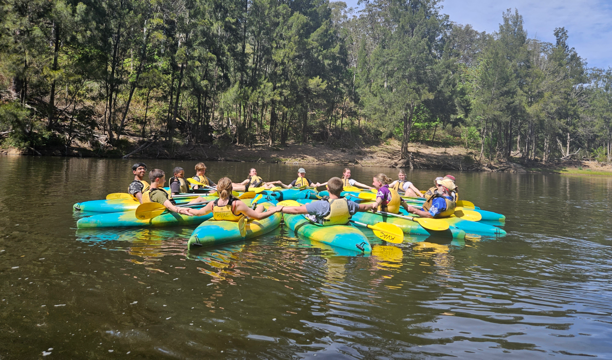 Multiple people on kayaks holding hands together in a circle peacefully on a calm river