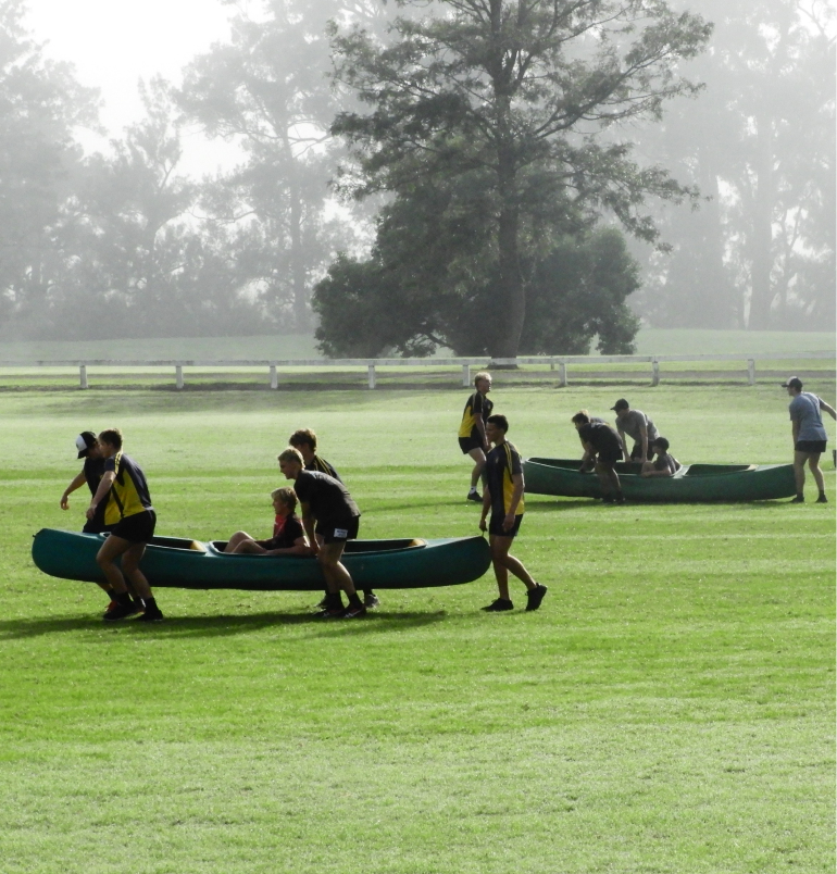 Teams working together carrying a person in a canoe over a grassed area