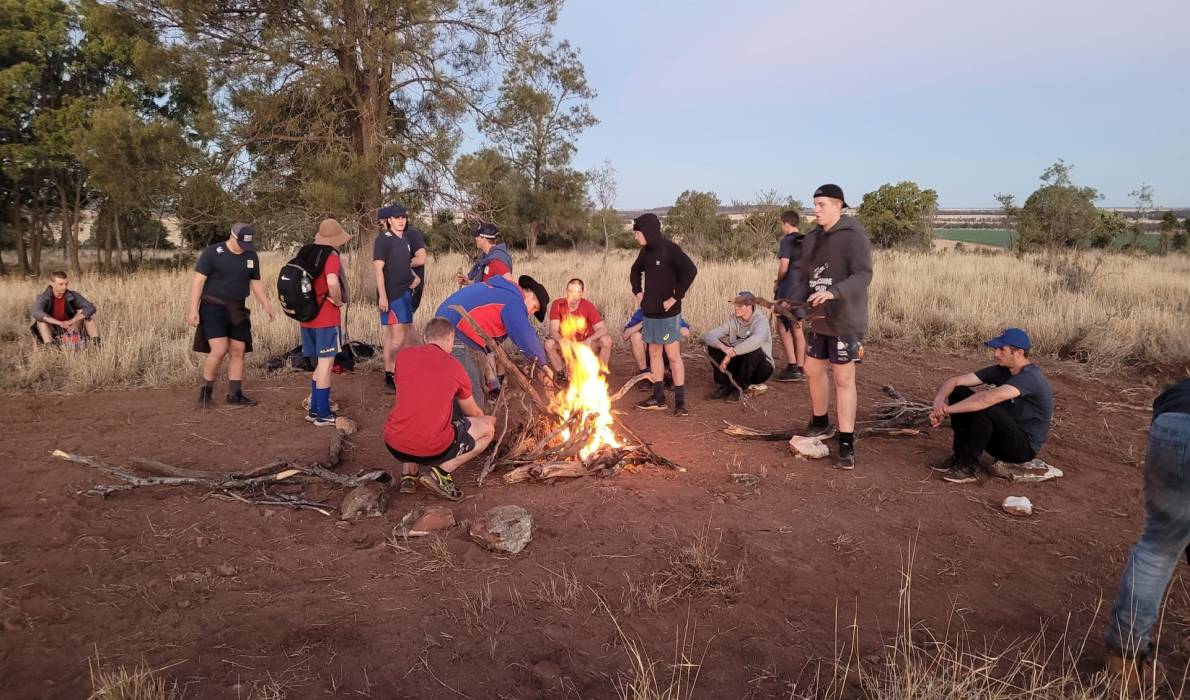 A group of people around a campfire at sunset, feeding the fire with sticks and wood